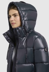 Giacca puffer blu scuro con colletto alto e cappuccio, caratterizzata da sezioni trapuntate lucide, cordini regolabili e un patch con logo.