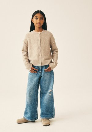 Jeune personne aux longs cheveux foncés porte un cardigan beige boutonné, un jean bleu large déchiré et des chaussures beiges, debout les mains près des poches.