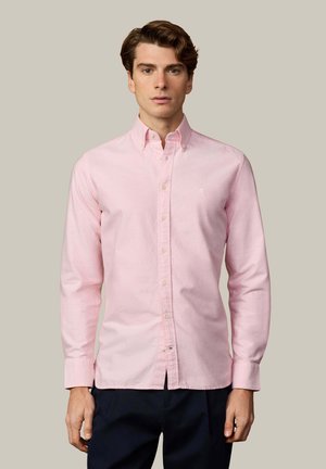Homme portant une chemise rose clair à boutons avec manches longues et un pantalon sombre, debout devant un fond beige uni.