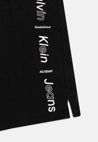 Czarne krótkie spodnie dresowe z białym napisem: "Calvin Klein Established" i "Jeans", z prostym rozcięciem po bokach.