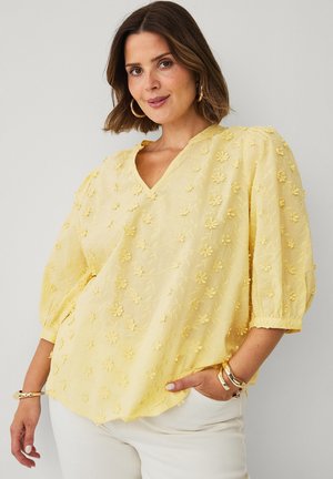 Kvinde iført en lysegul bluse med 3D blomsterapplikationer og puffede ærmer, kombineret med hvide bukser og gyldne hoopsøreringe.