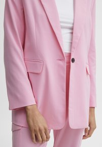 Pink blazer made of smooth fabric with a single button closure, notch lapels, and side pockets, paired with matching pink trousers.