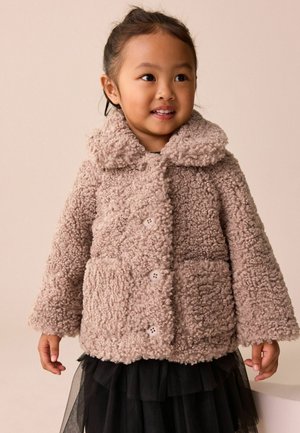 Ung pige smiler, iført en beige fluffy knapjakke med stor krave og et sort lagdelt skørt mod en ensfarvet baggrund.
