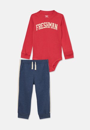 Roter Langarm-Bodysuit mit der Aufschrift "FRESHMAN" in weißer Schrift, kombiniert mit navyblauen Jogginghosen mit beigen seitlichen Streifen und einem Kordelzug.