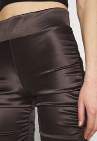Leggings en satin marron avec une taille haute, présentant des détails froncés sur les côtés et une texture lisse et réfléchissante.