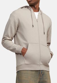 Haut à fermeture éclair beige clair avec capuche, poches avant, poignets côtelés et texture lisse. Comprend des cordons de serrage ajustables et un design simple.