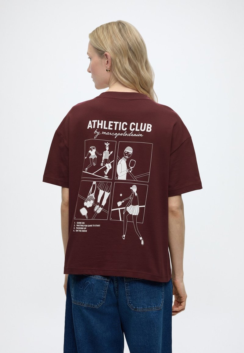 Žena s dlhými blond vlasmi má na sebe bordové tričko s bielymi grafikami a textom atletického klubu, v kombinácii s modrými džínsami, otočená chrbtom.