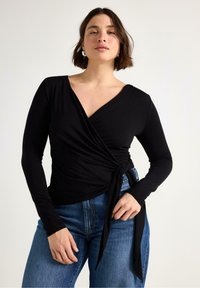 Svart långärmad omlottopp i mjukt tyg, med v-ringning och knytning vid midjan, kombinerad med blå högmidjade jeans.