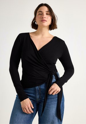 Top avvolgente a maniche lunghe nero realizzato in tessuto morbido, con scollo a V e dettaglio con nodo in vita, abbinato a jeans blu a vita alta.