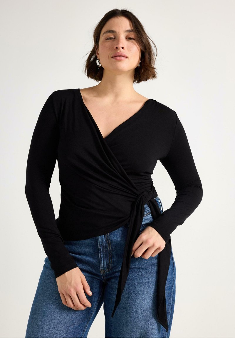 Svart långärmad omlottopp i mjukt tyg, med v-ringning och knytning vid midjan, kombinerad med blå högmidjade jeans.