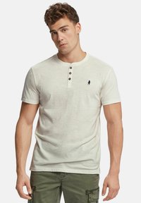 Gai beigā krāsā īsām piedurknēm henley krekls ar trim brūnām pogām uz apkakles; tam ir neliels tumši zils logo kreisajā krūšu pusē.
