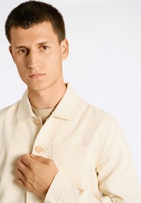 Ljust beige bomulljacka med knap stängning, krage och bröstficka. Slät textur, skräddarsydd passform. Modell som håller i kragen på vänster sida.