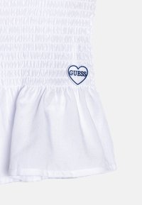 Weißes Baumwoll-Oberteil mit geraffter Struktur und Rüschensaum. Enthält ein blaues, herzförmiges "GUESS"-Logo auf der unteren linken Seite.