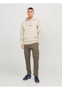 Sudadera beige con un bolsillo delantero y logotipo, combinada con pantalones cargo oliva que tienen bolsillos laterales. Zapatillas azul marino con detalles en azul claro.