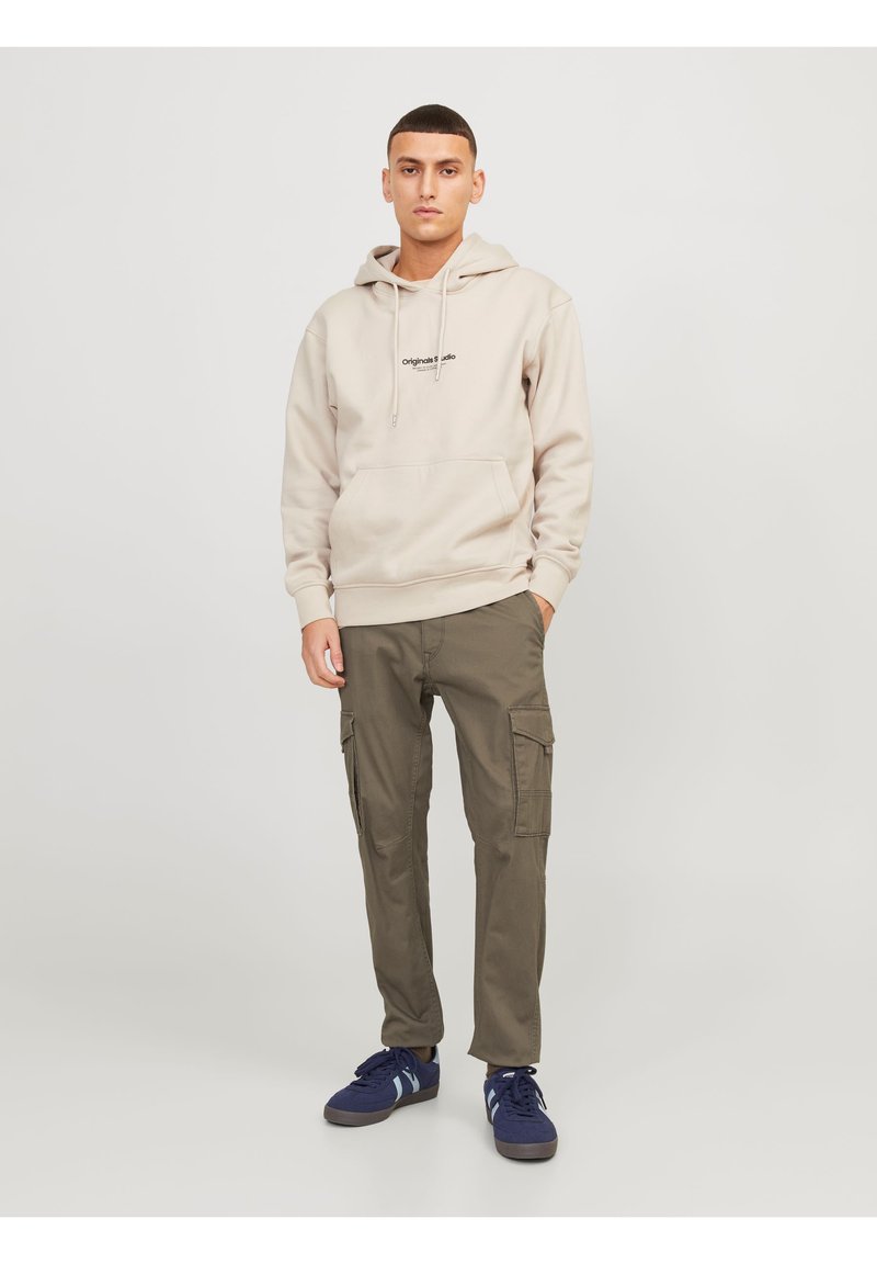 Sudadera beige con un bolsillo delantero y logotipo, combinada con pantalones cargo oliva que tienen bolsillos laterales. Zapatillas azul marino con detalles en azul claro.