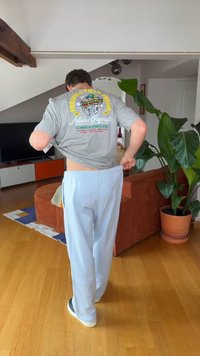 Man pulling up light blue pants, wearing a gray t-shirt with a colorful graphic on the back, standing in a living room with plants and furniture.