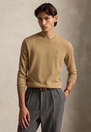 Jeune homme portant un pull beige à manches longues, un pantalon gris plissé avec une ceinture marron et une montre, debout avec une main dans la poche.