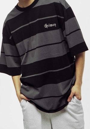 Personne portant un T-shirt ample à rayures horizontales noires et grises avec le texte « Grimey », mains dans les poches, associé à un pantalon gris clair.