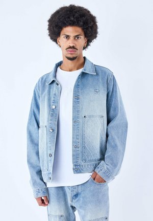 Giacca in denim azzurro chiaro con design a tasca trapuntata, bottoni argentati e vestibilità casual, indossata sopra una maglietta bianca.