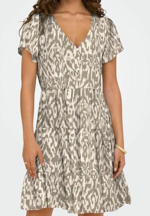 Vrouw die een beige-wit gemêleerde jurk met korte mouwen en knopen draagt, met een gelaagde rok en een gouden ketting en armband.