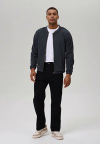 Jachetă bomber gri cu manșete și tiv ribb, purtată peste un tricou alb, asortată cu pantaloni negri și adidași ușori cu accente bej.