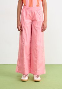 Pantalones de tiro ancho en rosa claro con una cintura elástica, que cuentan con franjas laterales y una textura suave. Combinados con zapatillas de estilo retro.