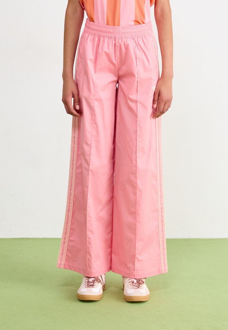 Pantalones de tiro ancho en rosa claro con una cintura elástica, que cuentan con franjas laterales y una textura suave. Combinados con zapatillas de estilo retro.