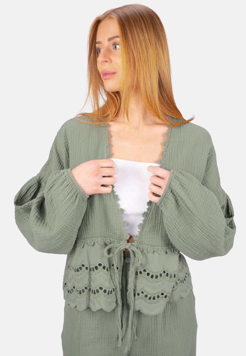 Hellgrüner strukturierter Cardigan mit Ballonärmel, Spitzenbesatz entlang des Halsausschnitts und Saums sowie Bindebändern in der Taille. Über einem weißen Oberteil getragen.