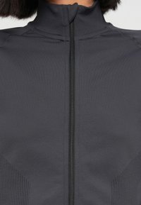 Gros plan sur une personne portant une veste gris foncé à fermeture éclair avec un col montant et des panneaux latéraux texturés visibles près de la fermeture éclair.