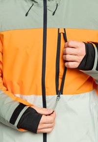 Chaqueta impermeable de color naranja brillante y verde claro con cremalleras negras, puños elásticos y un bolsillo con cremallera en el pecho.