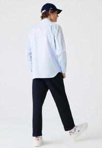 Lichtblauw overhemd met knopen, marineblauwe broek en witte sneakers met rode accenten. Hoed is marineblauw met een logo. Glad materiaal, relaxte pasvorm.