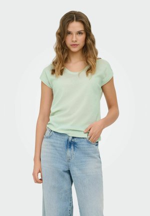 Vrouw met golvend haar draagt een lichtgroen topje met korte mouwen, ingestopt in een high-waist gebleekte blauwe spijkerbroek, staand tegen een effen achtergrond.