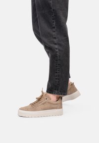 Sneaker marroni chiari con finitura testurizzata, punta rotonda, suola piatta bianca e design con lacci, abbinati a pantaloni di jeans scuri.