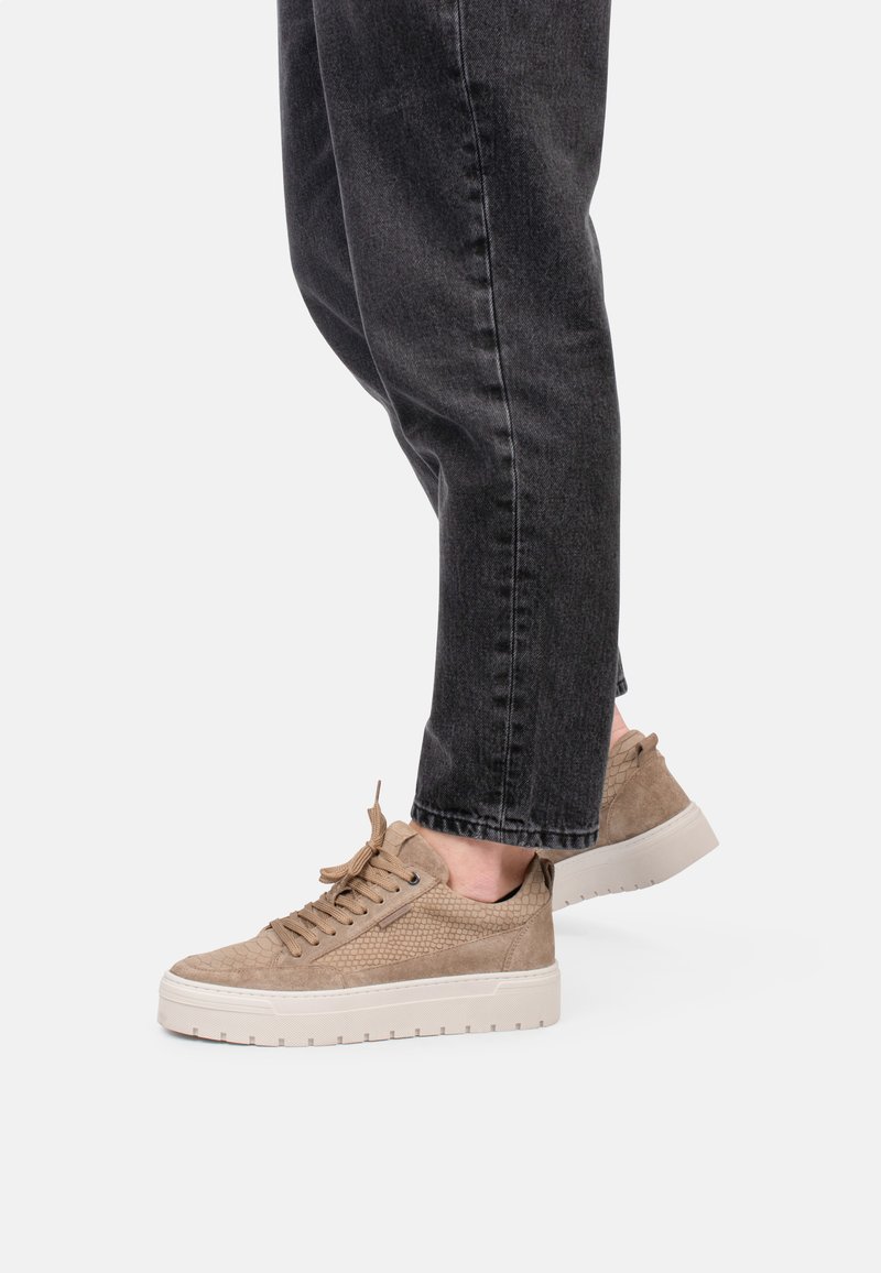 Sneaker marroni chiari con finitura testurizzata, punta rotonda, suola piatta bianca e design con lacci, abbinati a pantaloni di jeans scuri.