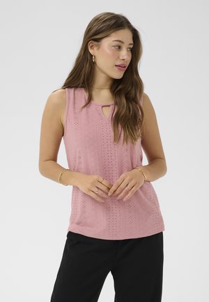 Frau mit langen Haaren, die ein ärmelloses, rosa strukturiertes Oberteil und schwarze Hosen trägt, seitlich blickend, die Hände locker vor der Taille verschränkt.