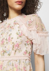 Robe rose brodée avec des motifs floraux, des manches transparentes à volants, et un tissu texturé, avec un détail de taille froncée.