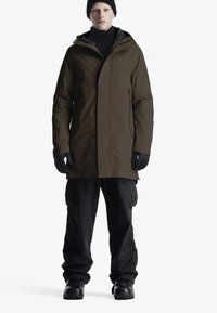 Persona che indossa un cappotto invernale marrone con cappuccio, berretto nero, guanti neri, pantaloni cargo neri e stivali neri, in piedi contro uno sfondo bianco.