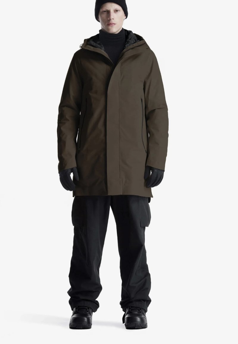 Persona che indossa un cappotto invernale marrone con cappuccio, berretto nero, guanti neri, pantaloni cargo neri e stivali neri, in piedi contro uno sfondo bianco.