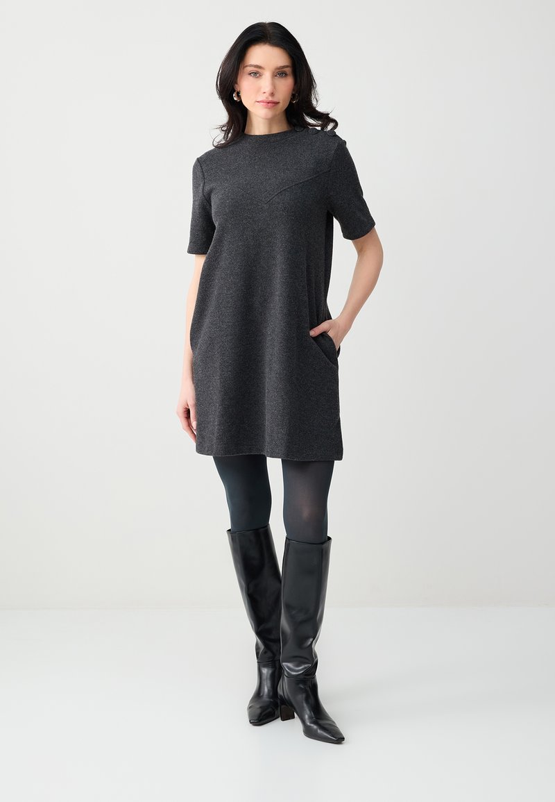 Femme portant une robe à manches courtes gris foncé, des collants noirs et des bottes noires montant jusqu'au genou, debout avec une main dans la poche.