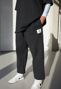 Dunkelgraue, lässig geschnittene Hose mit einem weißen quadratischen Jordan-Logo-Patch, kombiniert mit einem lockeren schwarzen T-Shirt und weißen Sneakers mit schwarzen Akzenten.