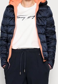 Giacca puffer blu navale con cappuccio arancione, pantaloni sportivi neri con tirante e una maglietta bianca con un logo blu navale. Texture liscia e trapuntata.