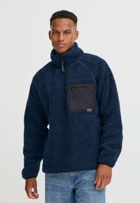Marineblauer Fleece-Pullover mit Halb-Zip-Kragen, ausgestattet mit einer schwarzen Reißverschlusstasche auf der Brust und strukturiertem Stoff, der für Wärme und Komfort sorgt.