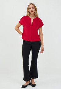 Blouse rouge à manches courtes avec un décolleté en V et un bouton décoratif en or à l'avant, associée à un pantalon noir évasé, complété par des ballerines noires.
