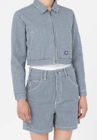 Kort blå och vit randig jacka med dragkedja, som har sidofickor, i kombination med matchande shorts. Båda har en texturerad kvalitet.