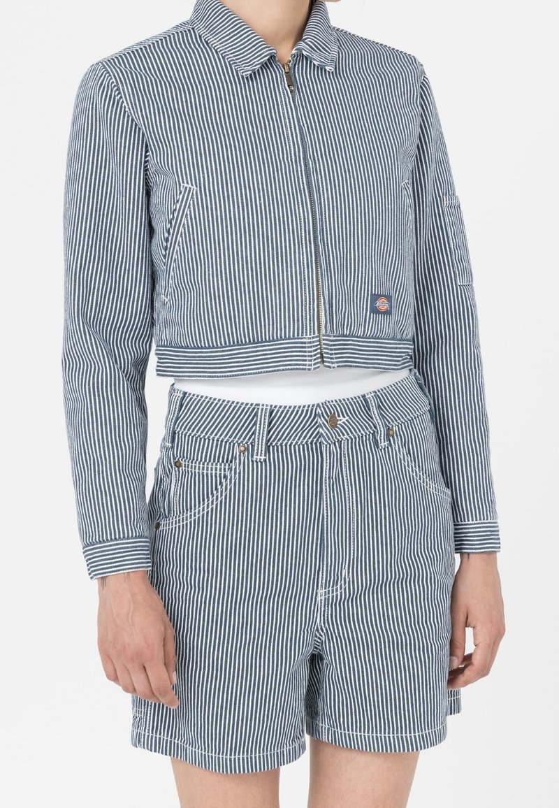 Kort blå och vit randig jacka med dragkedja, som har sidofickor, i kombination med matchande shorts. Båda har en texturerad kvalitet.