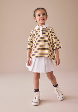 Young girl wearing a yellow and black striped polo shirt, white pleated skirt, dark socks, and white sneakers standing against a beige background.