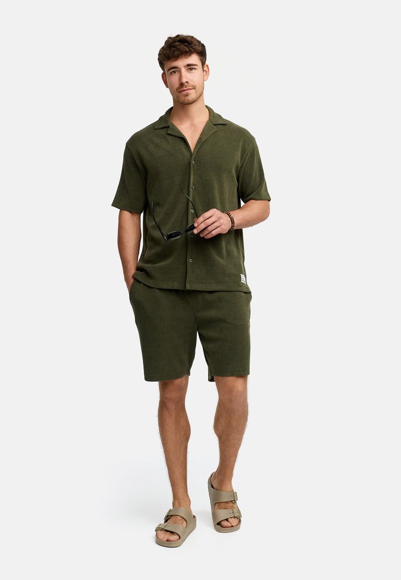 Camisa de botón de manga corta en color verde oliva con tejido texturizado, combinada con pantalones cortos a juego y sandalias beige. Sosteniendo unas gafas de sol en la mano.