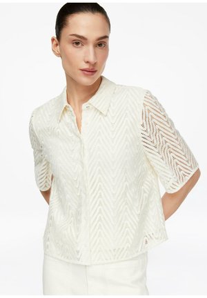 Frau trägt ein weißes Spitzenoberteil mit Chevron-Muster und Kragen, kurze Ärmel, kombiniert mit weißer Hose vor weißem Hintergrund.