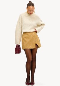 Suéter blanco esponjoso, falda amarillo mostaza con patrón floral, medias negras, bolso granate y zapatos de tacón rojo.