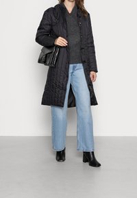 Abrigo largo acolchado negro con cierre de botones, suéter gris, jeans de pierna ancha en azul claro y botines negros de tacón. Incluye un bolso negro.